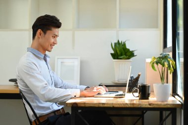Side view of smiling young asian male investor analyzing financial online data on laptop computer.