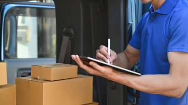 Cropped shot of delivery man in blue uniform using digital tablet next to open delivery van. Shipping and delivery service concept.