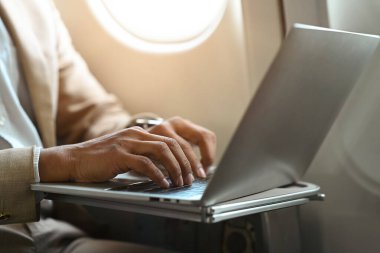 Cropped shot of businessman in suit typing, working on laptop while sitting in airplane cabin.