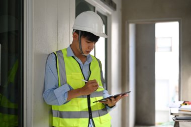 Asian male engineer in safety helmet using digital tablet, checking building construction progress.