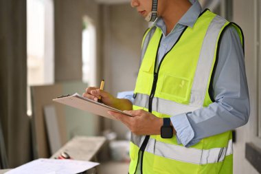 Engineer man in reflective jackets writing on clipboard, inspecting building construction site.