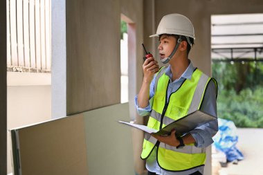 Asian male civil engineer manager standing at construction site and communicating with walkie talkie.