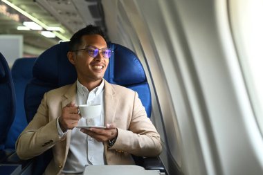 Successful businessman in elegant luxury suit drinking coffee and looking through window of the airplane during business travel.