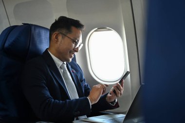 Side view of businessman passengers in black suit using smart phone while sitting on comfortable seat in airplane cabin.