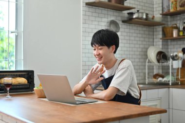 Cheerful young man freelancer checking email in morning, working online on laptop while sitting at wooden table in kitchen.