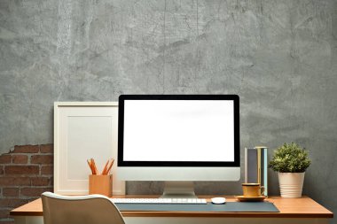 Blank computer monitor, picture frame, books, pencil holder and houseplant on wooden table against concrete wall. 