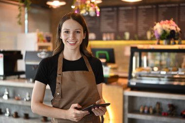 Half length of cheerful caucasian entrepreneur in apron standing in modern cafe interior and using digital tablet.
