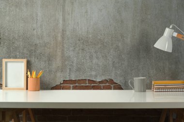 Stylish workplace with picture frame, books, pencil holder and lamp on wooden table against concrete wall. Copy space for your text.