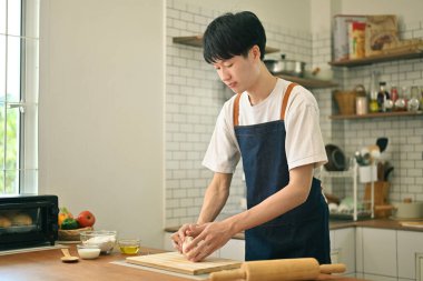Smiling young asian man wearing aprons hands kneading flour on wooden table, preparing homemade pastry in modern kitchen. 