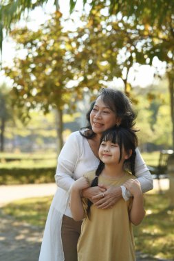 Happy middle age grandmother embracing grandchild while walking in public park surrounded by green trees at sunlight morning.