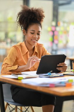 Smiling African American female employee searching online information on digital tablet, sitting at her workplace.