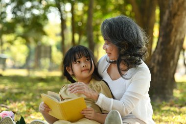 Loving grandmother reading fairytale to adorable little grandchild while sitting together in nature park. Family and love concept.