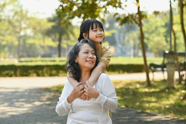 Happy moments with grandmother and little granddaughter walking in the park surrounded by green trees at sunlight morning.