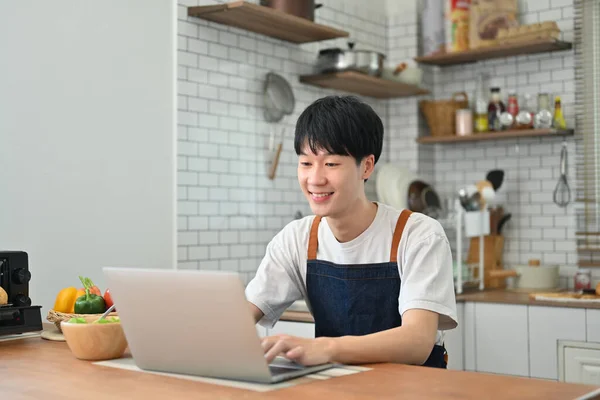 Smiling young asian man freelancer using laptop, working remote job from home while sitting in cozy kitchen interior.