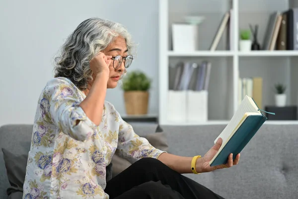 Elderly woman trying to read book having difficulties seeing text because of vision problems.