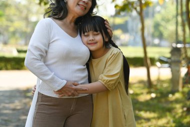 Cute little grandchild hugging her mature grandmother, standing in public park. Family and love concept.