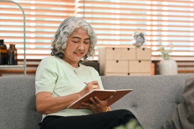 Relaxed middle aged woman sitting on couch and reading interesting book, enjoying leisure weekend time at home.