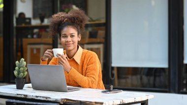 Charming young female freelancer drinking coffee and using laptop in an outdoor cafe.
