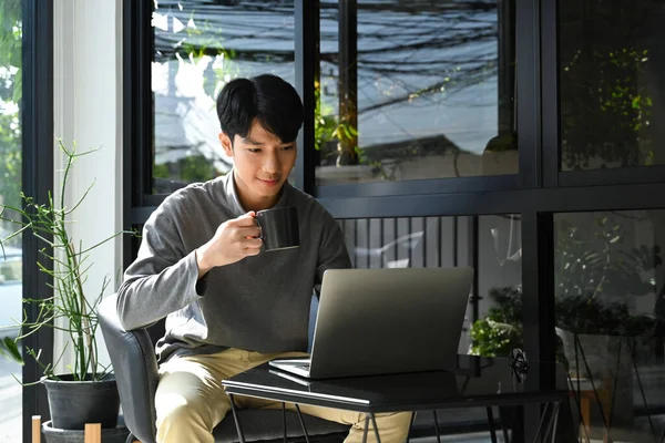 Pleasant man in warm clothes drinking coffee and checking email on laptop computer.