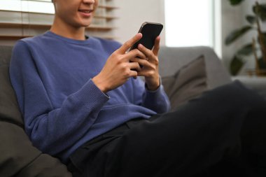Cropped shot of man typing text message, chatting online on mobile phone while sitting in couch at home.