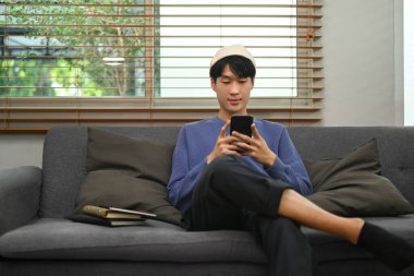 Hipster man typing text message, chatting online on mobile phone while sitting in couch at home.