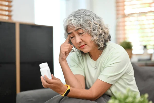 Worried middle aged woman taking medicine while sitting on couch at home. Elderly healthcare, medication concept.