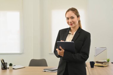 Gorgeous businesswoman with digital tablet in hands standing in corporate office and smiling to camera.