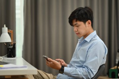 Handsome man freelancer sitting in modern home office and chatting online in social network with smart phone.