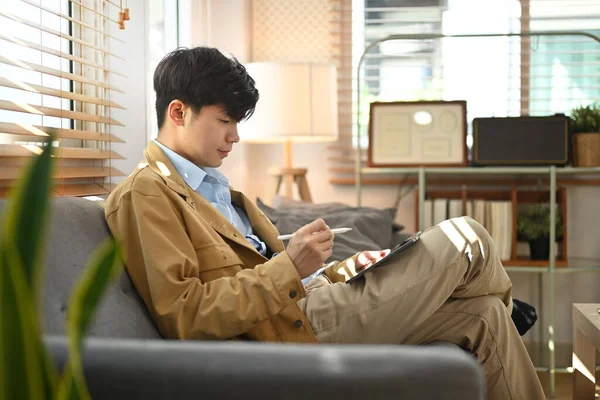 Side view of asian male freelancer working online, checking email on tablet while sitting in living room.