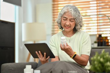 Mature woman holding medical pills bottle having online consultation with doctor on digital tablet. Elderly healthcare.