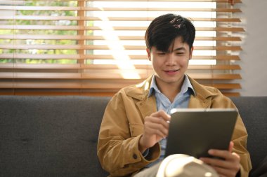 Relaxed millennial man working online or checking social app on digital tablet while sitting in living room.