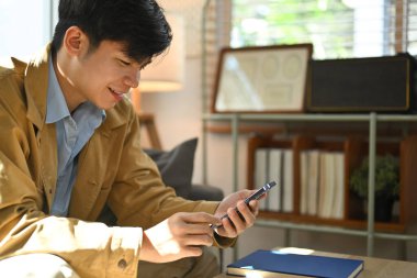 Pleased asian man sitting in bright living room and using smart phone. People, technology and lifestyle concept.