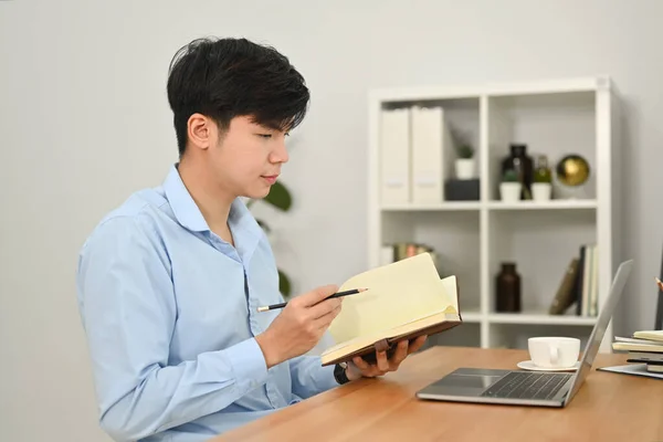 Young businessman looking at laptop screen and making important notes on notepad.