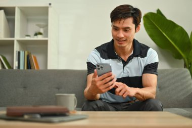 Smiling asian man resting on comfortable couch and typing text message on mobile phone.