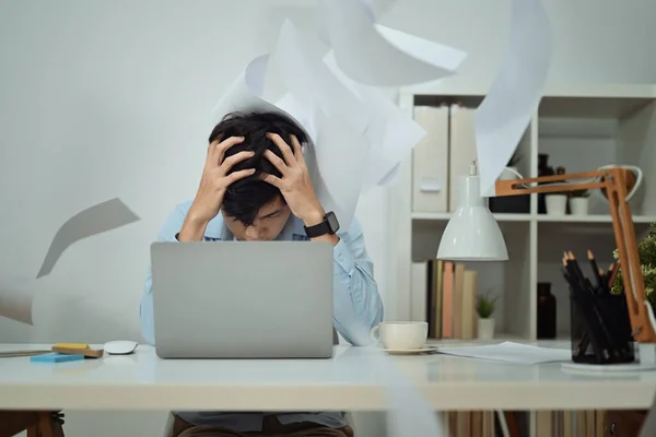 Stressed man office worker sitting at desk with falling paperwork, feeling distressed anxious with work deadline.