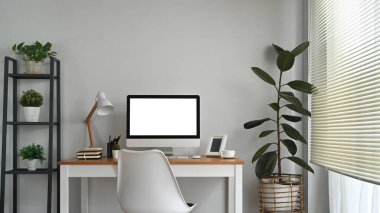 Bright home office interior with blank computer screen, lamp and supplies on wooden desk.