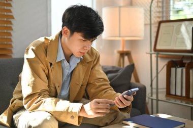 Millennial asian man relaxing on couch in cozy living room and enjoy browsing wireless internet on mobile phone.