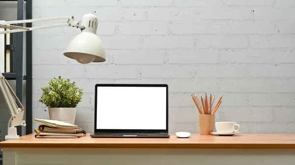 Laptop computer, potted plant, lamp and supplies on wooden desk against brick wall. Blank screen for advertise design.