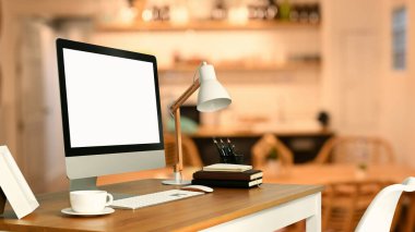 White blank screen computer monitor, lamp, coffee cup and books on table with blurred kitchen on background.