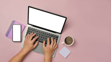 Overhead view of woman hands typing on laptop computer with smart phone and coffee cup on pink background.