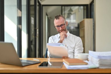 Portrait of experienced business man read document paper report front of laptop at hid workplace.
