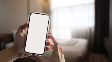 Hands holding smart phone with blurred light bedroom interior on background. White screen for graphic display montage.