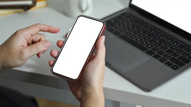 Man holding smartphone white screen while sitting in front of laptop at workplace. Blank screen for advertise webpage or text. 