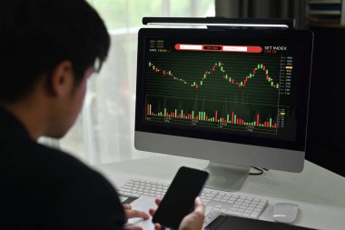 Side view of young male financial analyst using mobile phone and monitoring stocks data candle charts on screen.