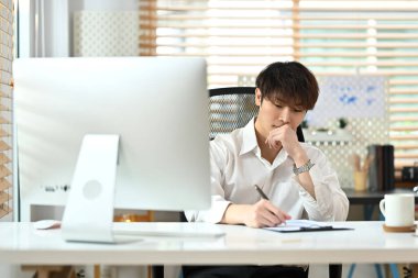 Handsome asian businessman wearing white shirt and making notes on the documents at his workplace.