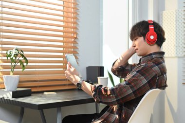 Image of young hipster man in stylish casual wear listening to music in headphone and using digital tablet at home office.