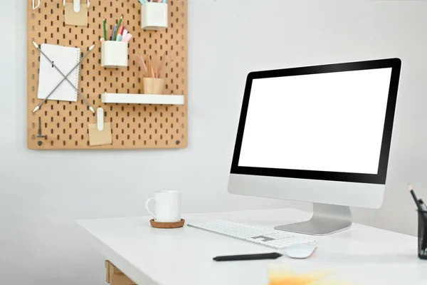 Blank computer screen and supplies on white table with pegboard on wall. Empty screen for advertising design.