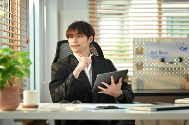 Thoughtful businessman holding digital tablet and looking through window, pondering idea or developing business strategy.