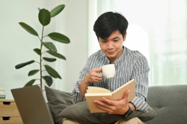 Joyful millennial man drinking coffee and checking his working schedule plan in cozy living room.