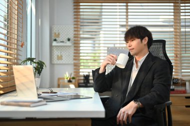 Pleasant asian male worker in formal clothes drinking coffee and reading business email on laptop computer.